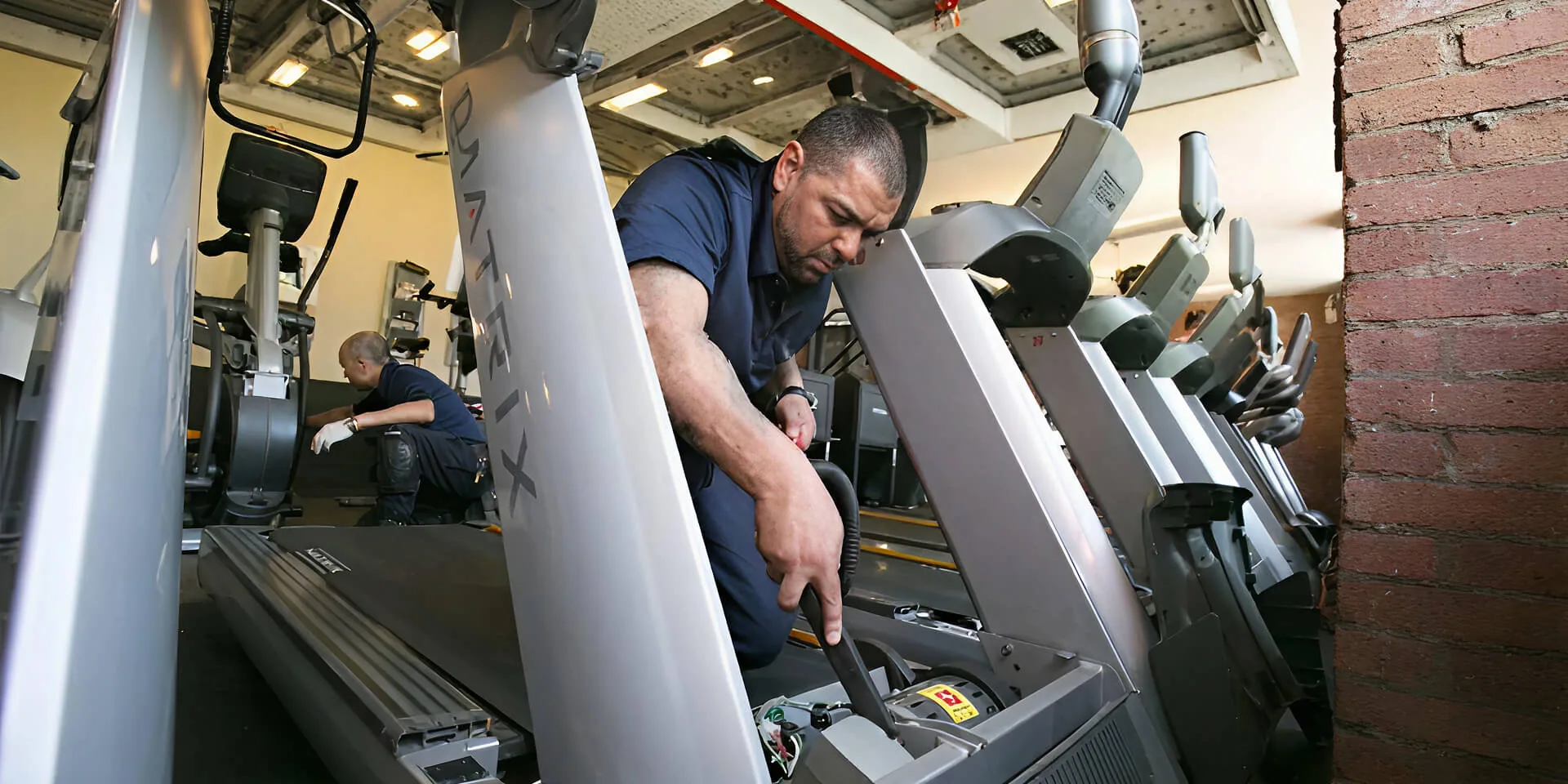 Technician servicing an opened treadmill console while a second technician works behind him